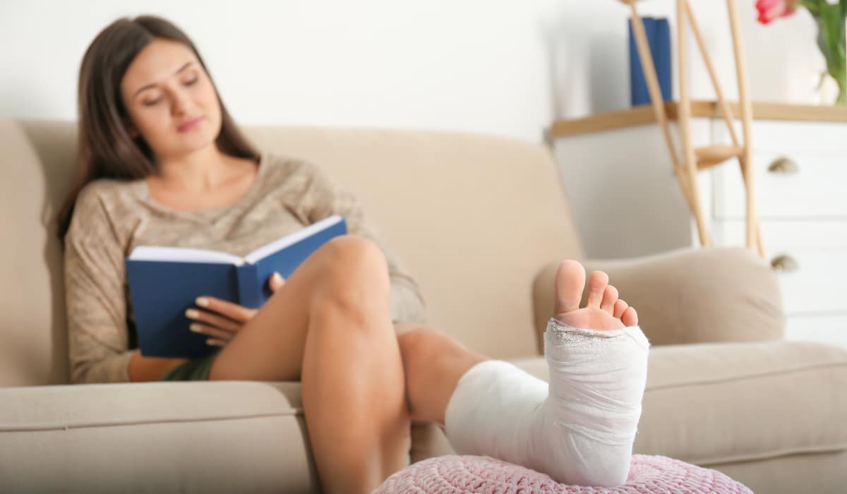 Young woman with broken leg in cast reading book while sitting on sofa at home